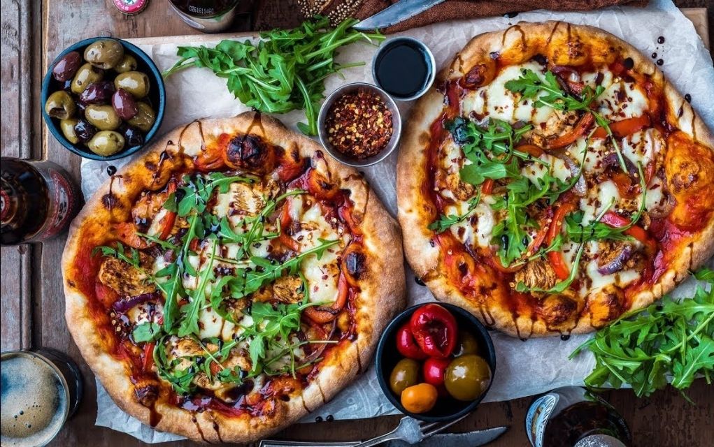 two pizzas and deli food on a table viewed from above