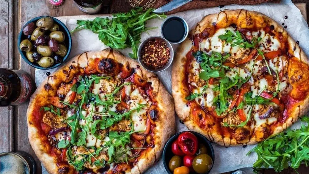 two pizzas and deli food on a table viewed from above