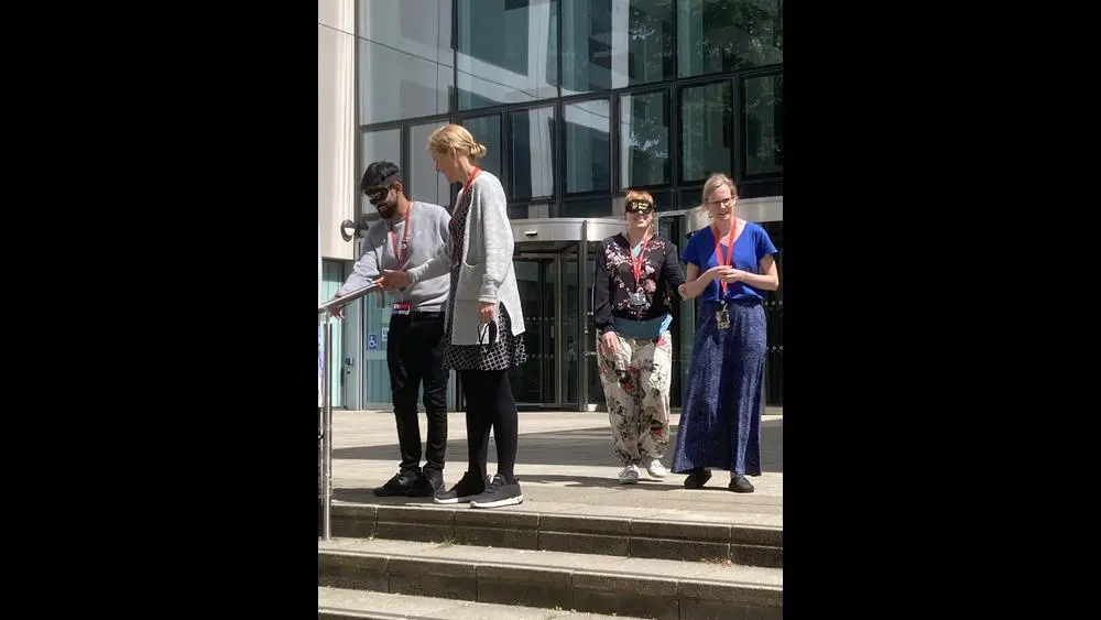 Members of staff wearing blindfolds being guided down the steps outside the Spark by other members of staff