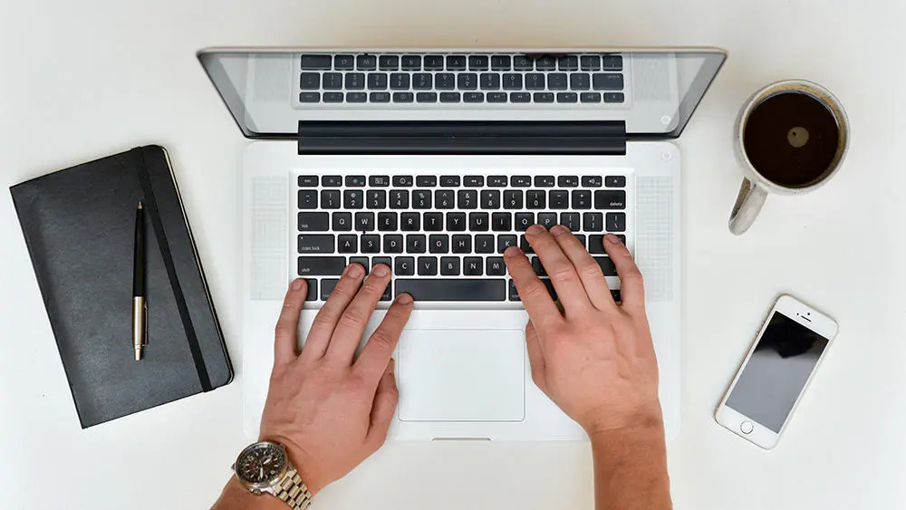 A person typing on a laptop with coffee and notebook